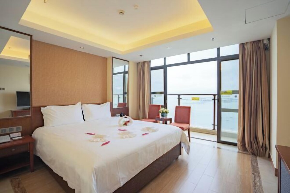 A hotel room with a large bed, a desk, a chair, a TV, and a view of the sea.
