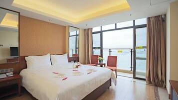 A hotel room with a large bed, a desk, a chair, a TV, and a view of the sea.