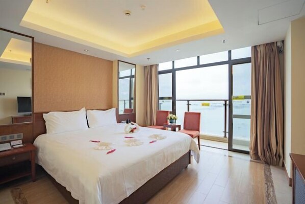 A hotel room with a large bed, a desk, a chair, a TV, and a view of the sea.