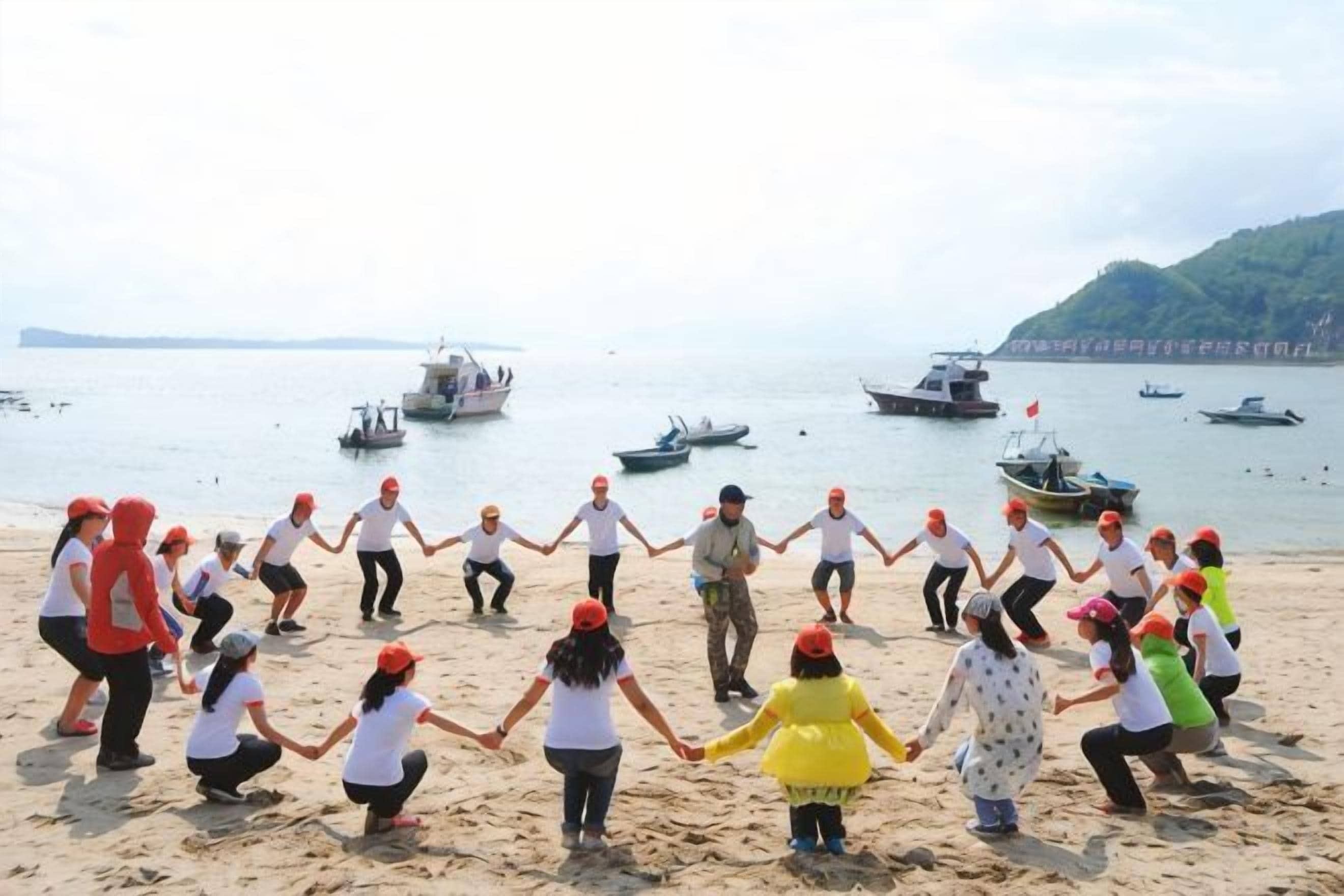 Un grupo de personas tomadas de la mano en una playa arenosa con barcos en el agua.