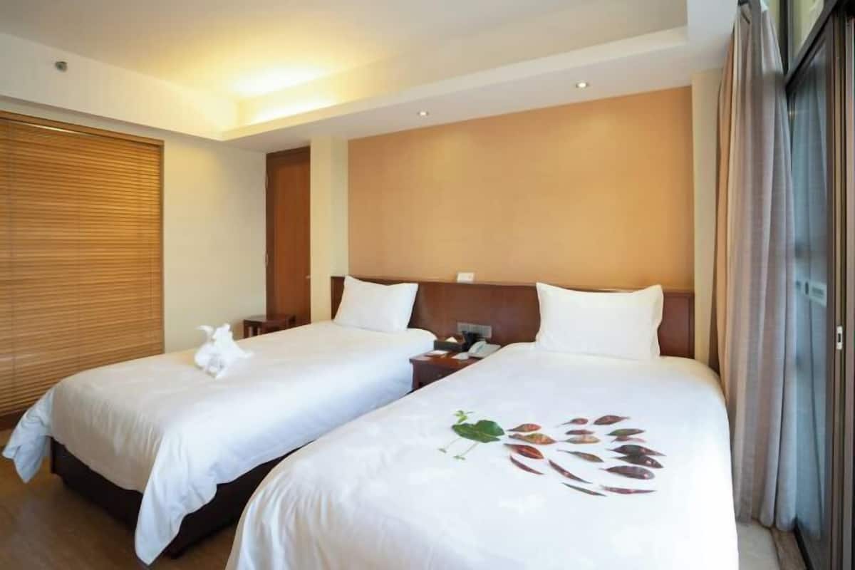Habitación de hotel con dos camas, cabecera de madera, mesita de noche con lámpara y ventana con persianas.