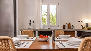 Un coin repas avec une table en bois dressée pour quatre personnes, entourée de chaises en osier. Un saladier de tomates est posé sur la table, ainsi que deux bols blancs et un couteau.