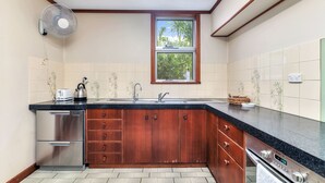 A kitchen with a built-in oven, a stainless steel refrigerator, wooden cabinets, a black countertop, a window with a view of palm trees, and a wall-mounted fan. - Tropical Crush | 4 Beds, 2 Bath | Pool (Fannie Bay)