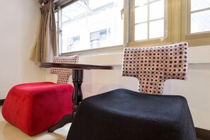 A room with a window, a table, two ottomans, and a patterned chair.