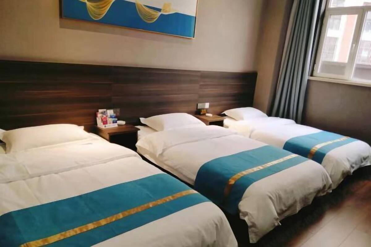 Habitación de hotel con tres camas, cabecera de madera, mesita de noche con lámpara y un cuadro en la pared.