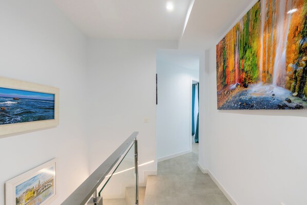A hallway with a staircase, a framed painting of a coastal scene, and a large abstract painting on the wall. - Corazón de Triana. Terrace & private pool (Sevilla)