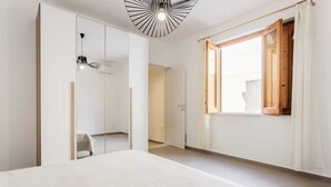 Een slaapkamer met een bed, een kledingkast, een spiegel, een raam met gordijnen en een plafondlamp.