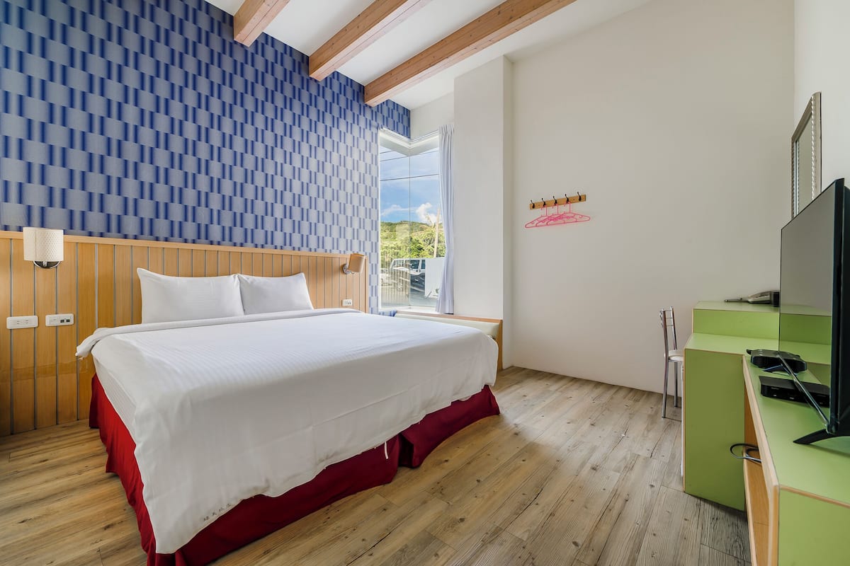 A bedroom with a large bed, wooden floor, a TV, and a window with a view of greenery.