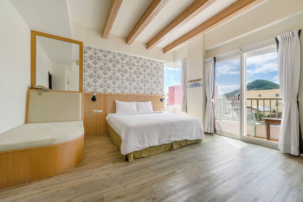 A bedroom with a bed, a bedside table, a mirror, and a view of the outside.