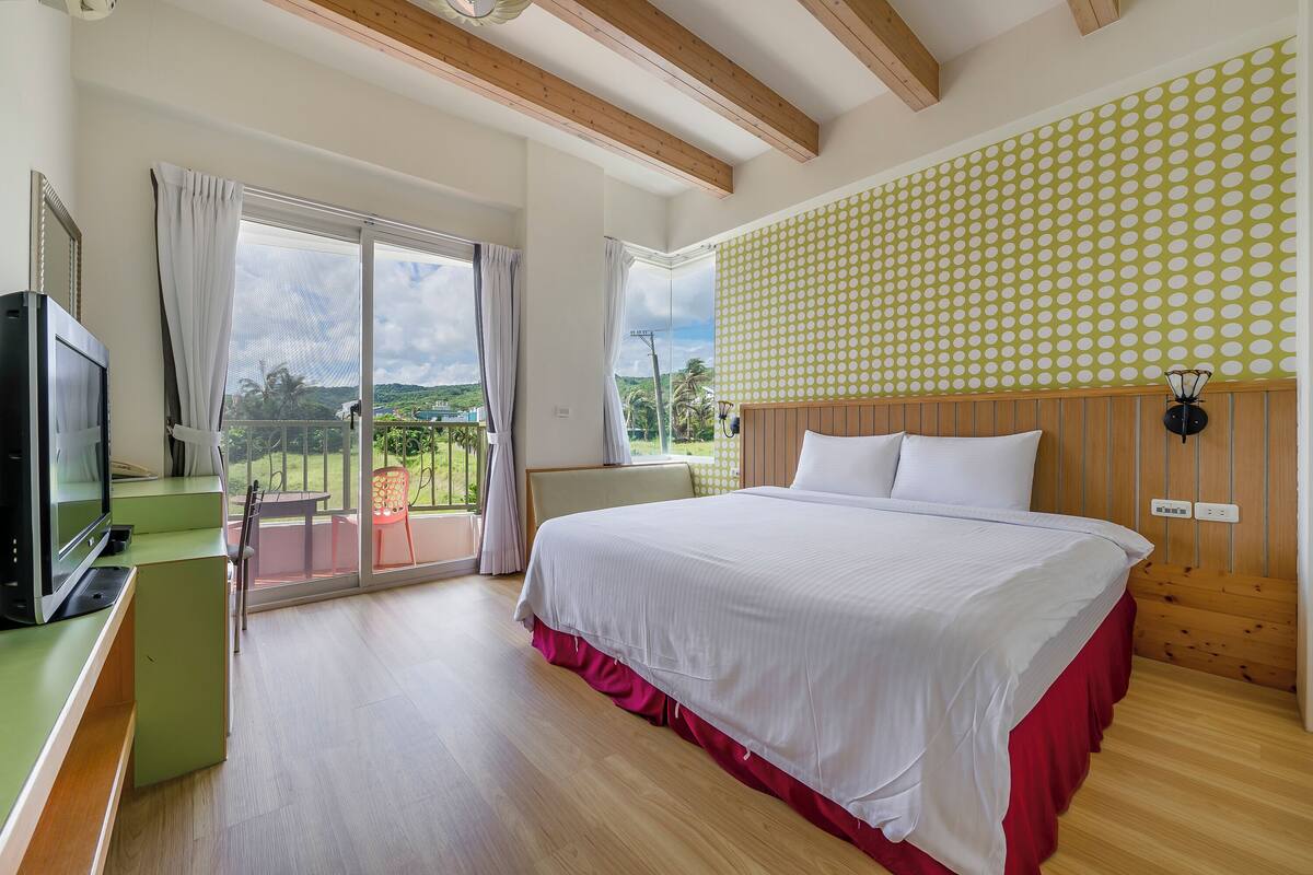 A bedroom with a large bed, a TV, a balcony with a view, and a patterned wall.