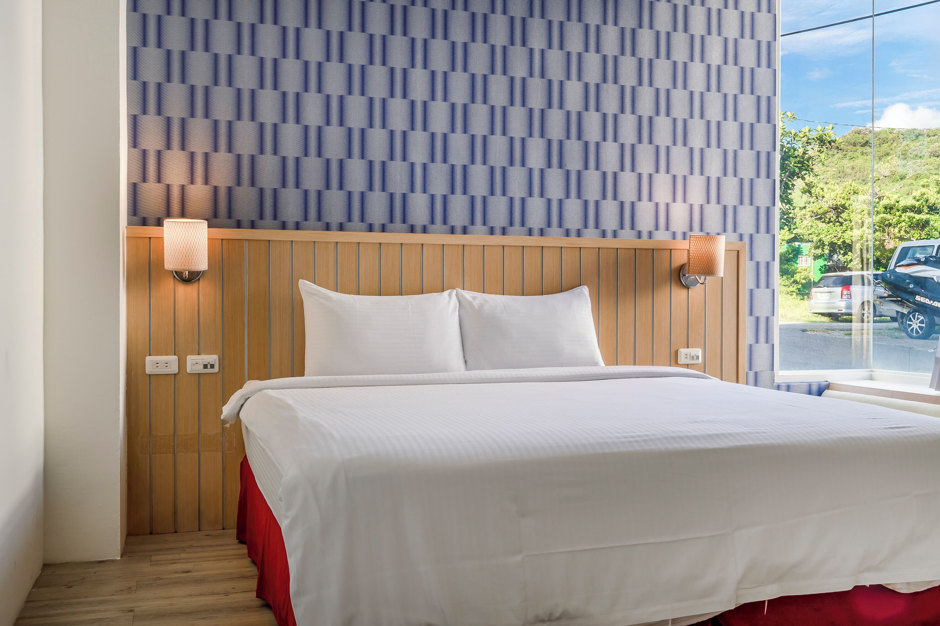 A hotel room with a bed, wooden headboard, two wall-mounted lamps, and a window with a view of a parking area.