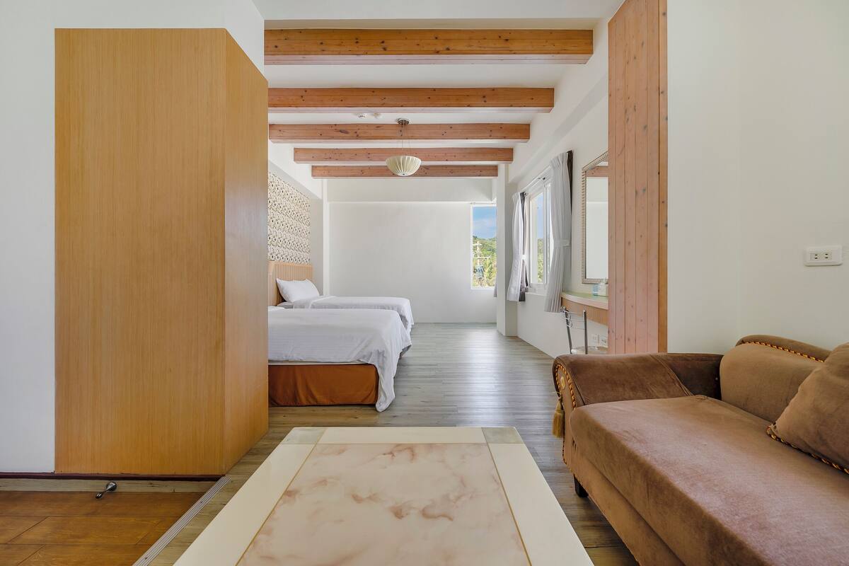 A bedroom with a bed, a wooden wardrobe, a brown sofa, and a view of the outdoors.