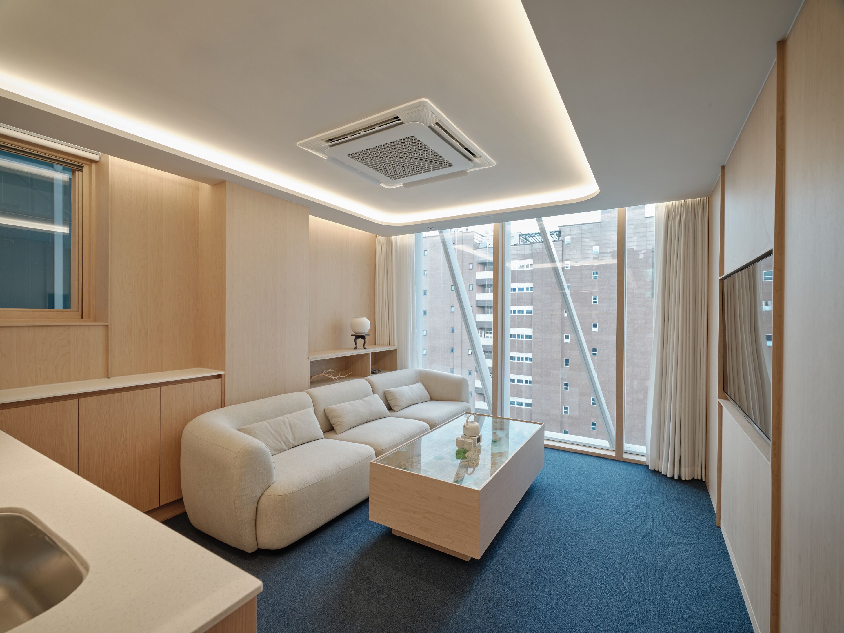 A modern hotel room with a sofa, a glass coffee table, a built-in sink, and a large window offering a city view.