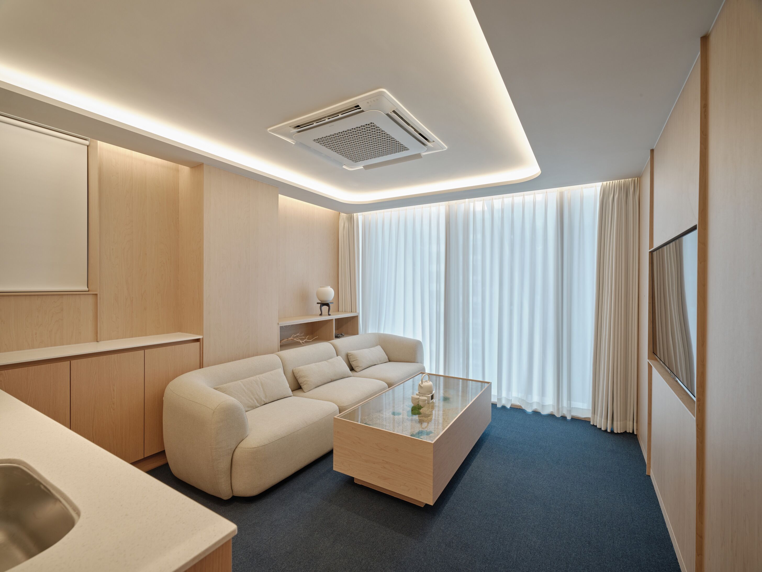 A modern hotel room with a sofa, a glass coffee table, a built-in shelf, and a large window with curtains.