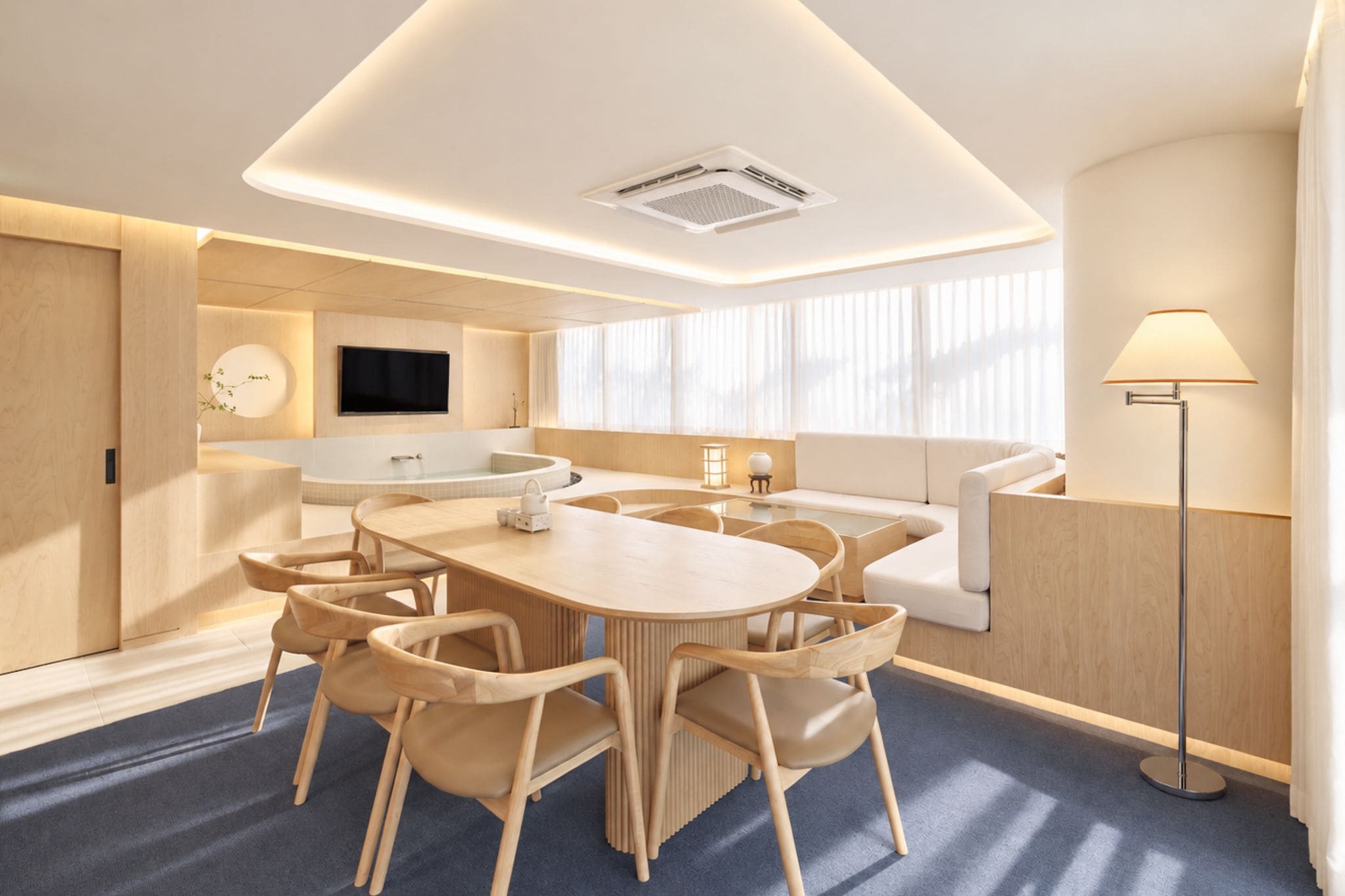 A modern hotel room with a round dining table, chairs, a sofa, a flat-screen TV, and a floor lamp.