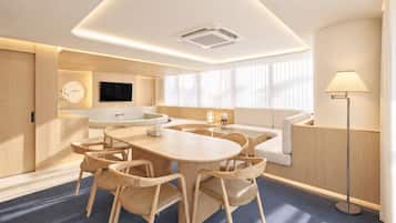 A modern hotel room with a round dining table, chairs, a sofa, a flat-screen TV, and a floor lamp.
