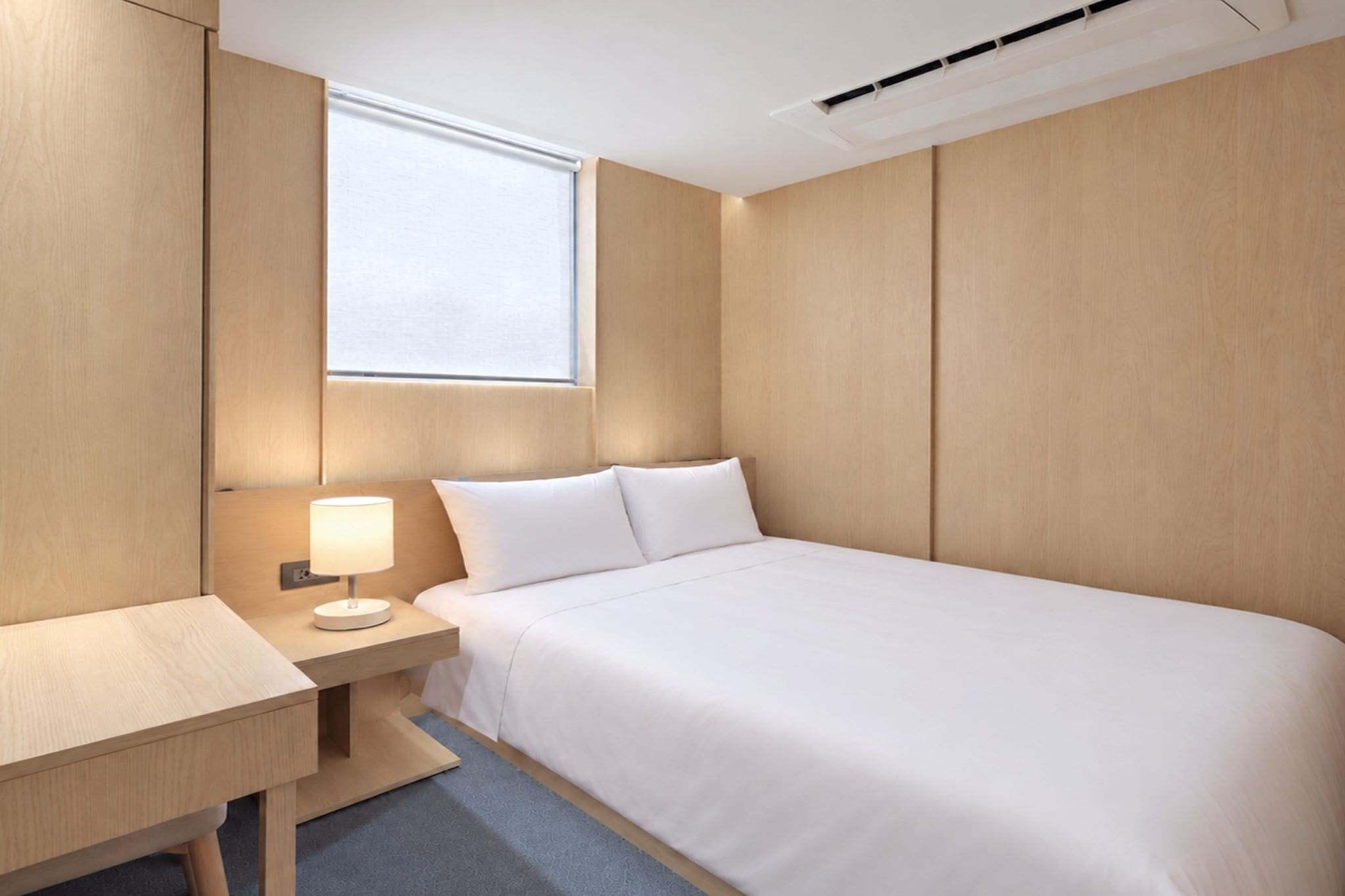 A hotel room with a bed, bedside table, lamp, and a window.