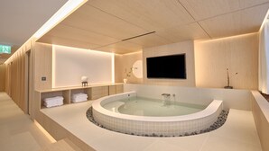 A modern spa room with a large bathtub, wooden paneling, a flat-screen TV, and neatly folded towels. - UH Suite The Gangnam (Seoul)