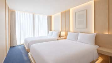 A hotel room with two beds, a large window with curtains, a bedside table with a lamp, and a framed picture on the wall.