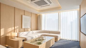 A modern hotel room with a sectional sofa, a glass coffee table, a bedside table with a lamp, and a large window with sheer curtains.