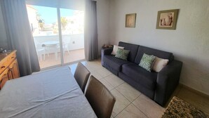 A room with a bed, a sofa, a dining table, and a balcony with a view of buildings and palm trees. - Las Floritas 3 (Playa de Las Americas)