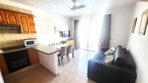 A modern kitchen with a microwave, oven, and wooden cabinets. There is a dining area with chairs and a table. A dark-colored sofa is in the living area. - Las Floritas 3 (Playa de Las Americas)
