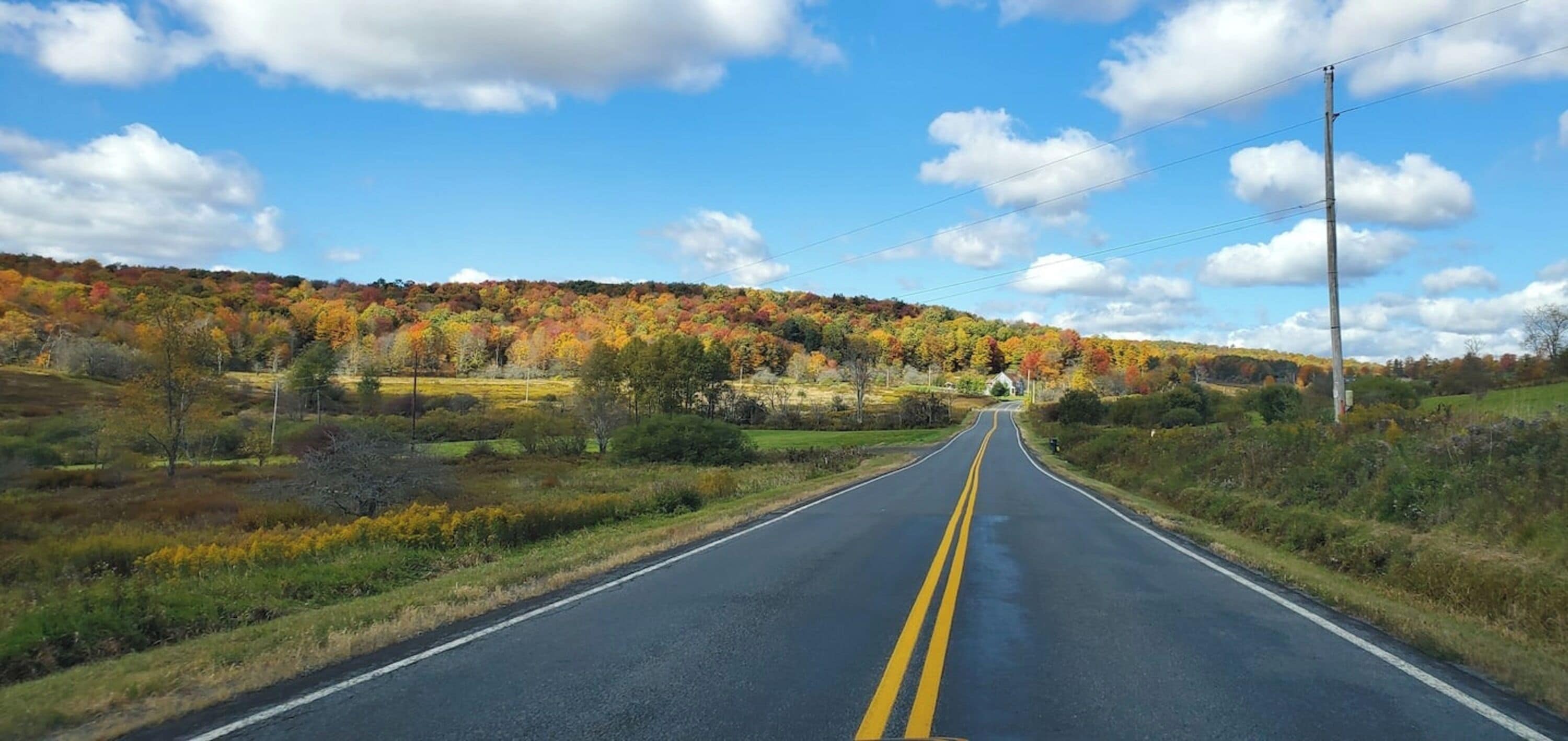 A two-lane road with a yellow center line, surrounded by autumn-colored trees and a clear blue sky.
