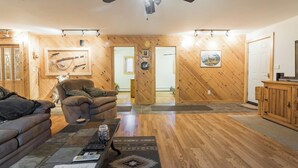 Ein geräumiges Wohnzimmer mit Holzwänden, Deckenventilator und Kamin. Es gibt ein Sofa, einen Sessel und einen Couchtisch mit Fernbedienung und einem Buch.