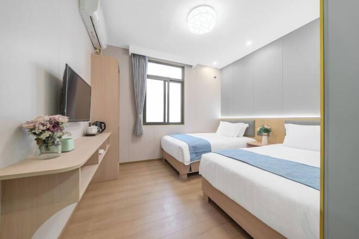 Habitación de hotel con cama, mesita de noche, televisor, ventana y un pequeño escritorio con un florero.