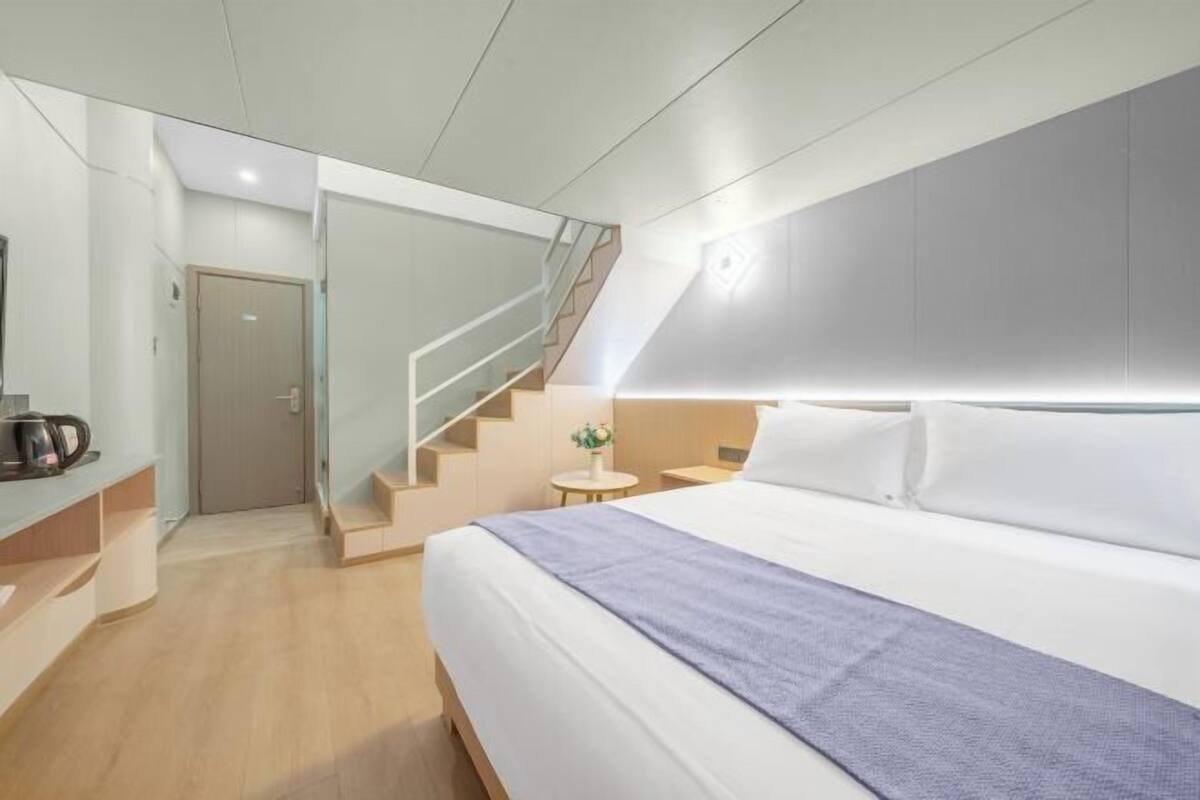 Una habitación de hotel moderna con una cama grande, una mesita con una lámpara, una cafetera y una escalera.