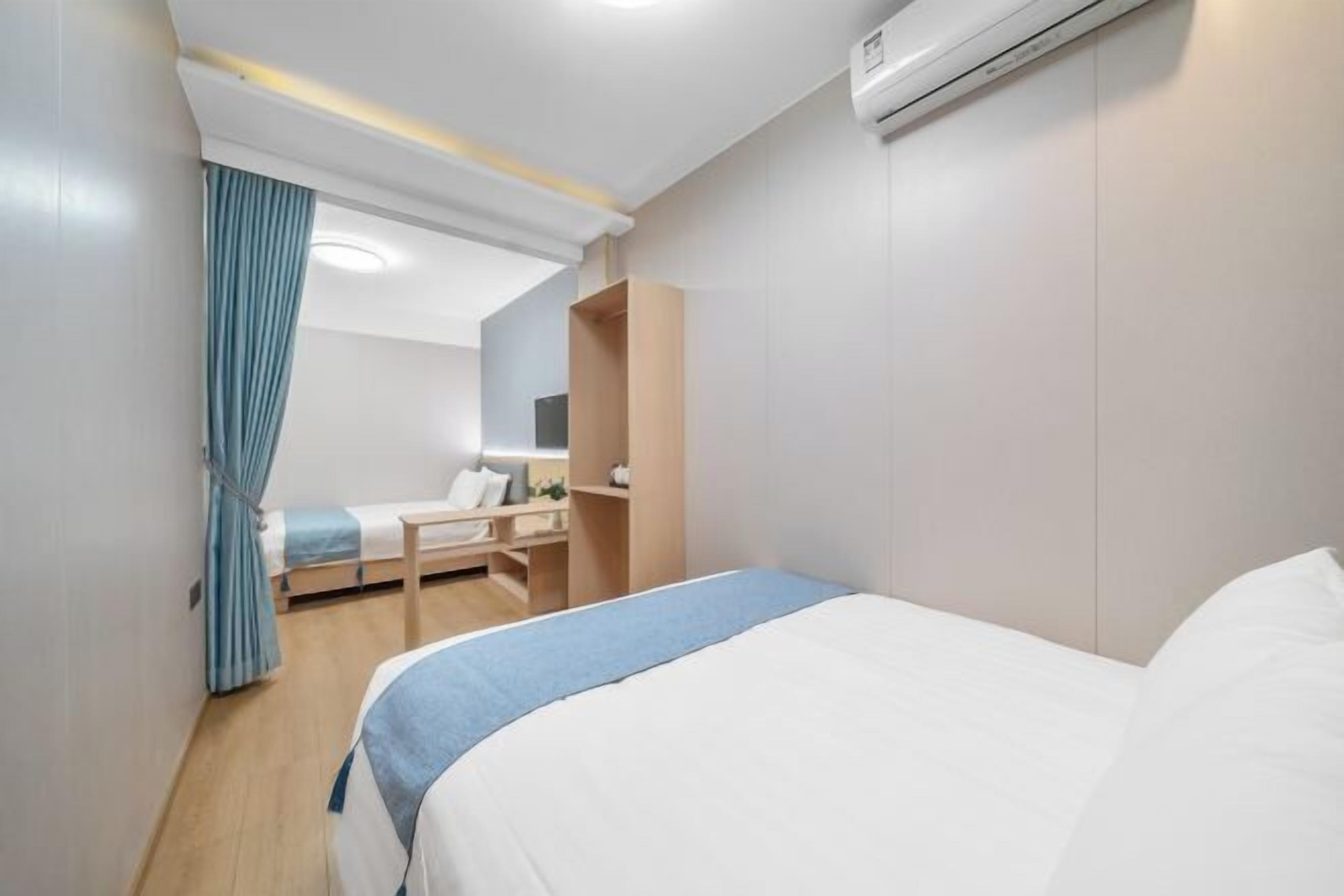 A hotel room with a bed, a desk, a chair, a wardrobe, and an air conditioning unit.