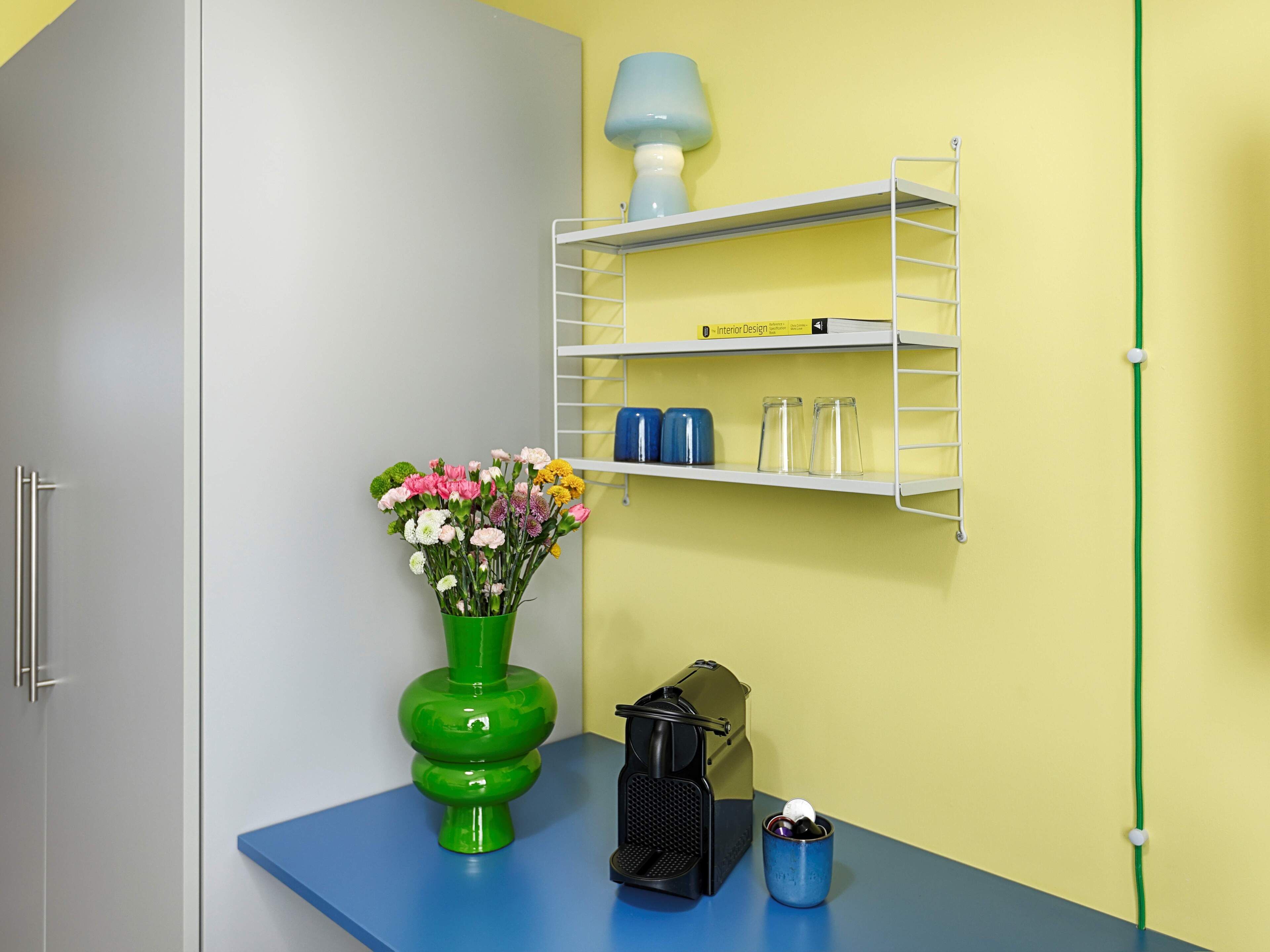 A room with a blue table, a green vase with flowers, a black radio, and a shelf with kitchen items.