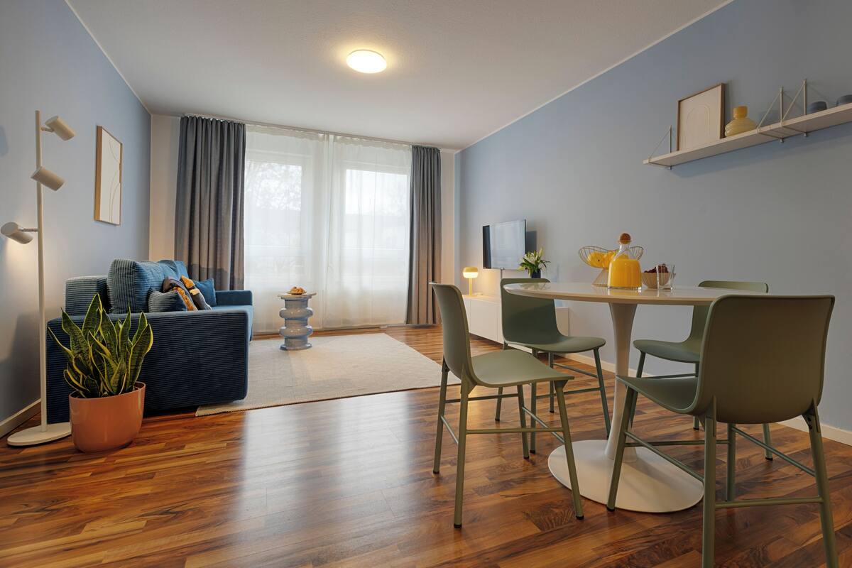 A modern living room with a sofa, a dining table with chairs, a potted plant, and a TV on the wall.