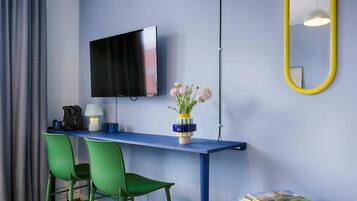 Una habitación con una televisión de pantalla plana en la pared, un escritorio con dos sillas verdes, un jarrón con flores y un espejo con marco amarillo.