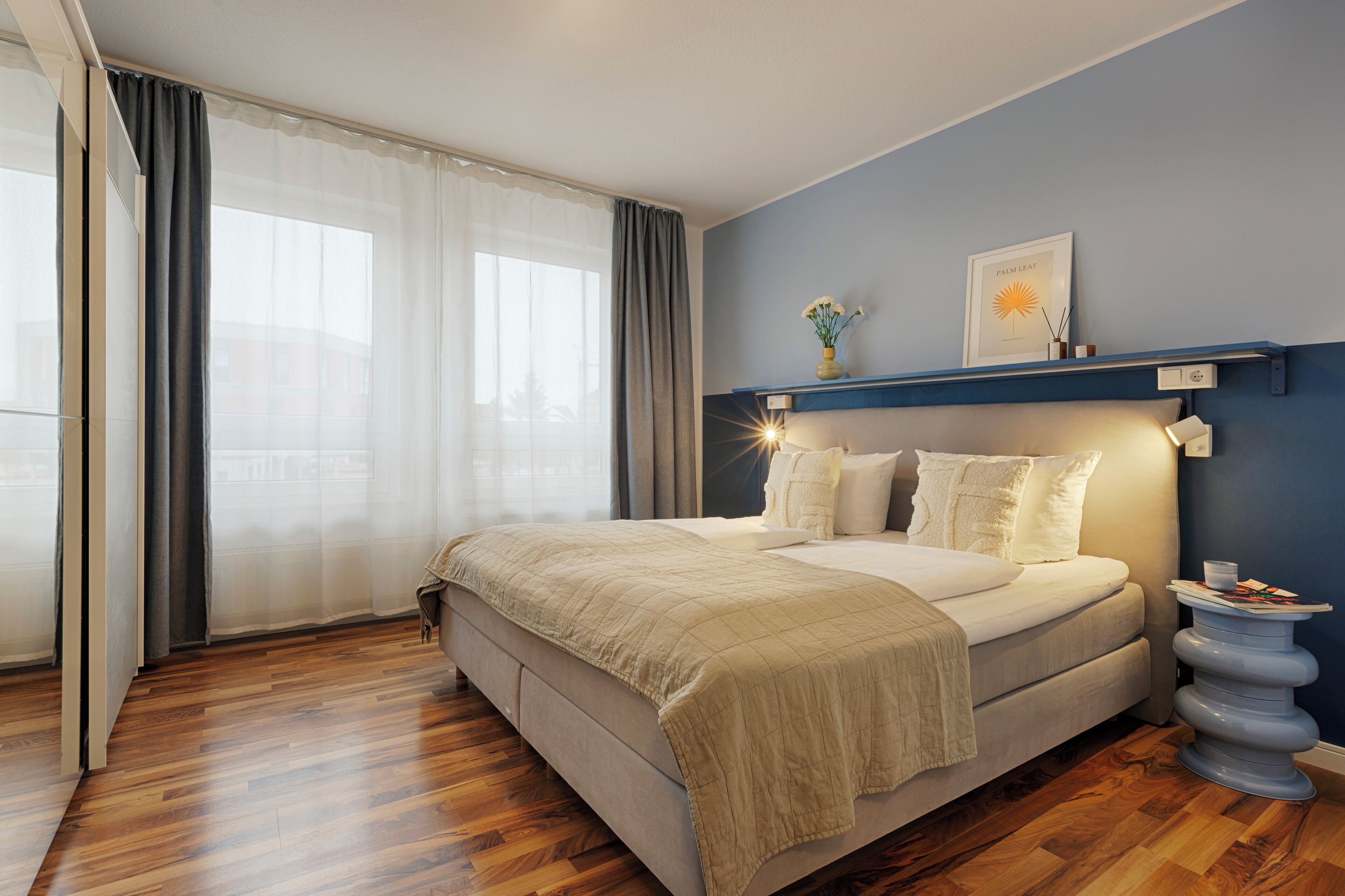 Un dormitorio con una cama grande, una mesita de noche, un estante de pared con adornos y una ventana con cortinas traslúcidas.