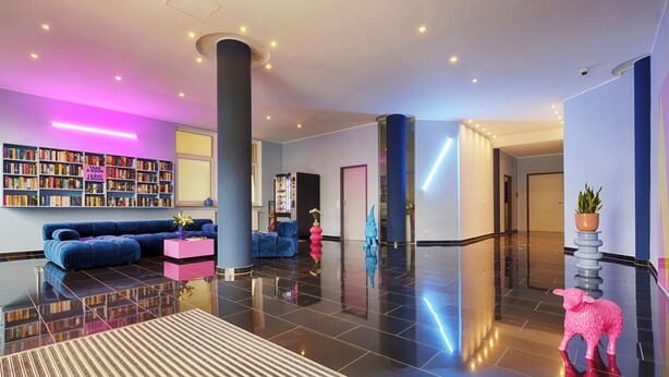 A modern lobby with a blue sofa, a pink lamb sculpture, and a bookshelf filled with books. - homie hotel max apart (Fuerth)