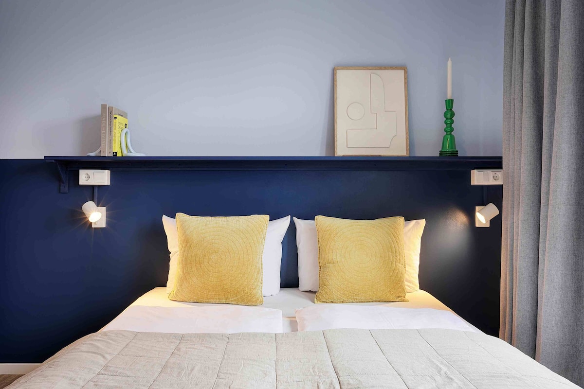 A neatly made bed with two pillows, a shelf with books and a candle, and a wall-mounted light fixture.