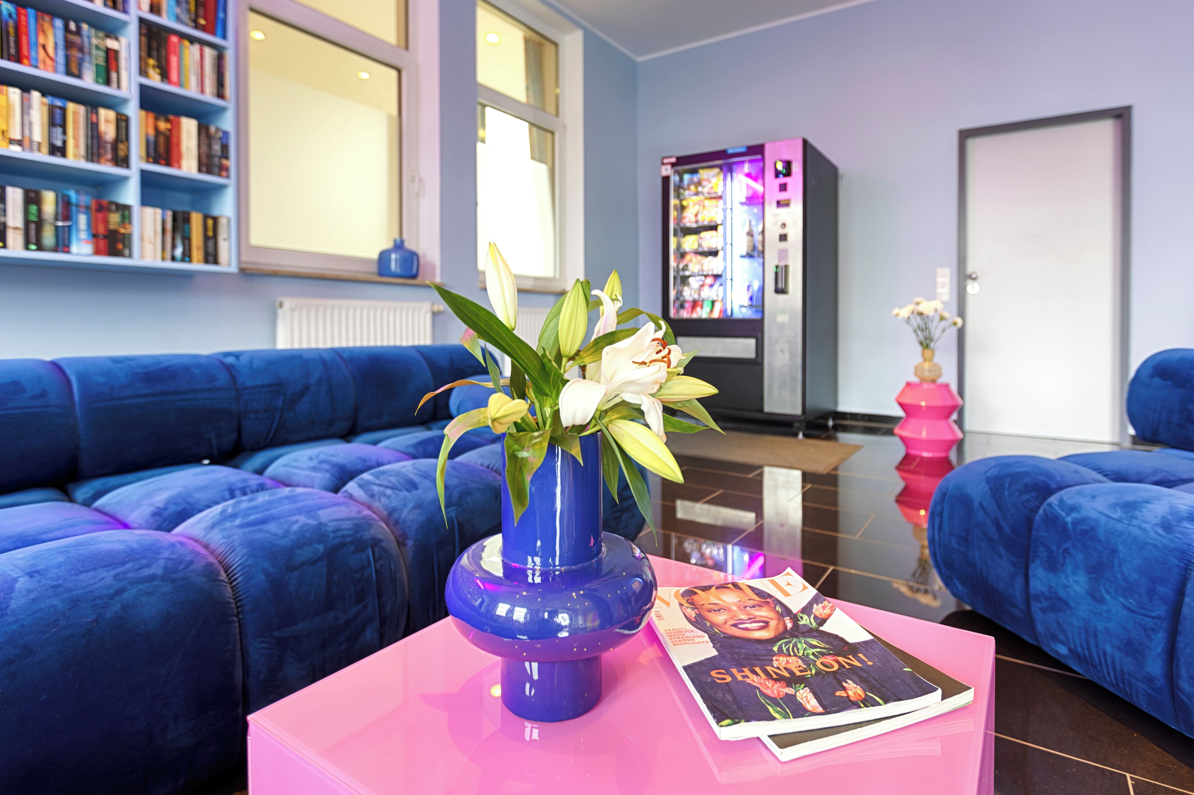 A modern living room with a blue sofa set, a pink coffee table with a vase and magazine, and a bookshelf filled with books.