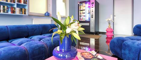 Un salon moderne avec un canapé bleu, une table basse rose ornée d’un vase et d’un magazine, et une bibliothèque remplie de livres.