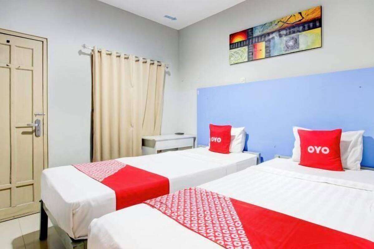 Une chambre d’hôtel avec deux lits, une tête de lit blanche, un oreiller rouge orné du mot « OYO », un rideau beige et un tableau accroché au mur.