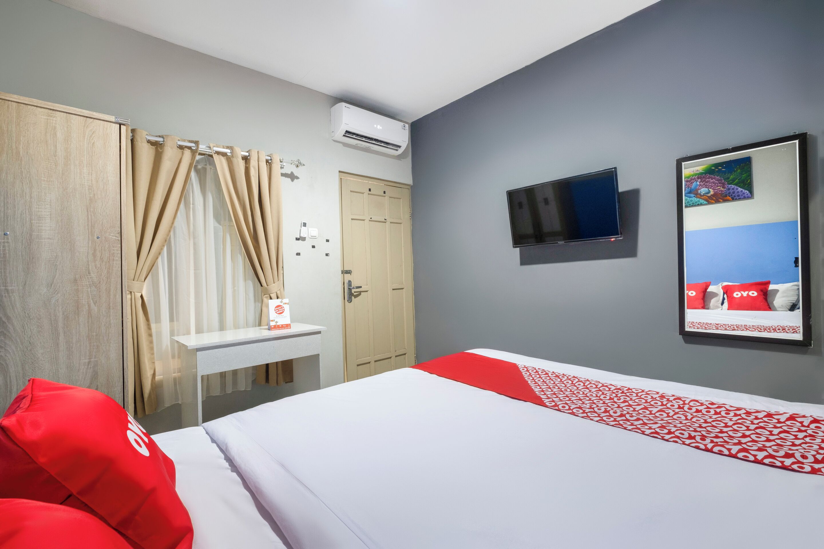 A hotel room with a bed, a desk, a wardrobe, an air conditioning unit, a television, and a painting on the wall.