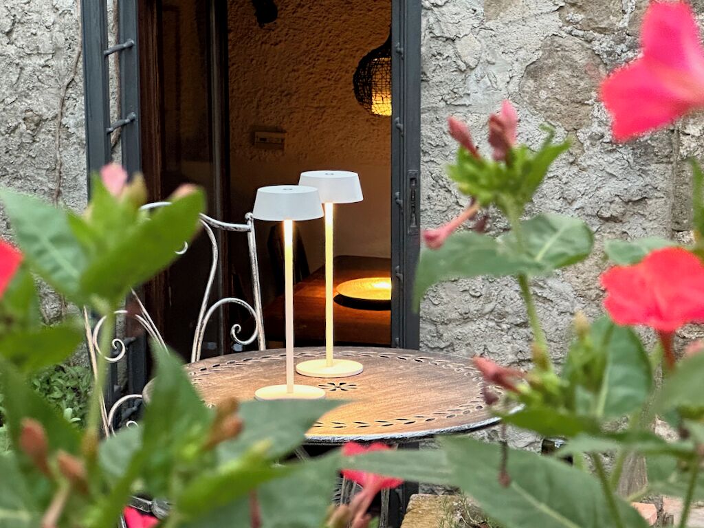 Zona d'seients a l'exterior amb estil rústic, amb una taula rodona i dues làmpades, envoltada de vegetació i una paret de pedra.