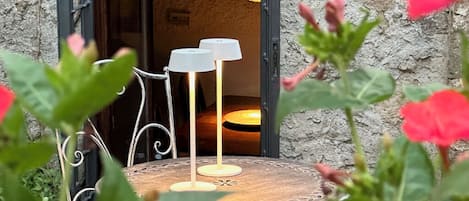 A rustic outdoor seating area with a round table and two lamps, surrounded by greenery and a stone wall.
