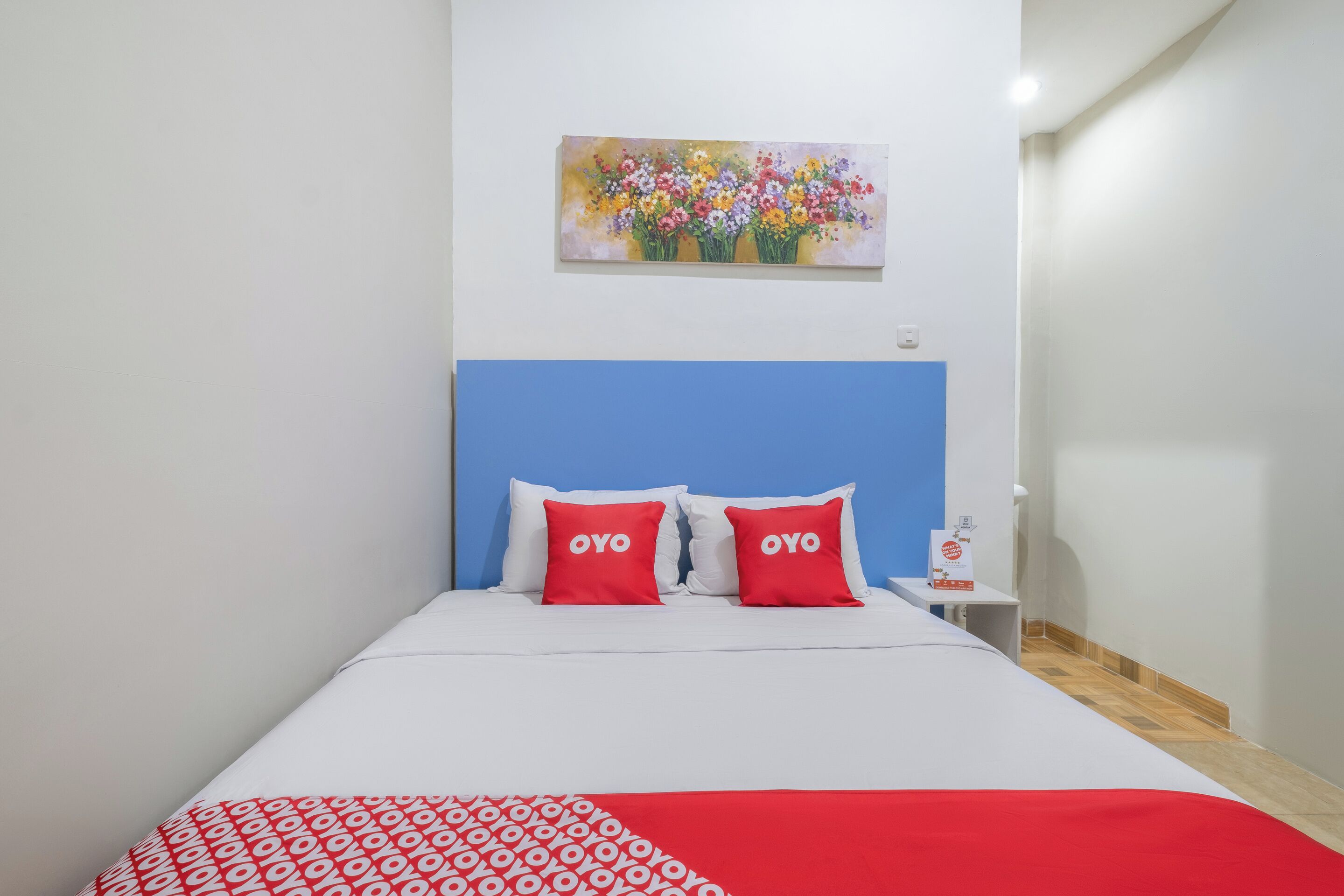 A bed with a blue headboard, white bedding, and two red pillows with "OYO" text. A colorful floral painting hangs above the bed.