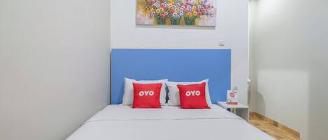 A bed with a blue headboard, white bedding, and two red pillows with "OYO" text. A colorful floral painting hangs above the bed.