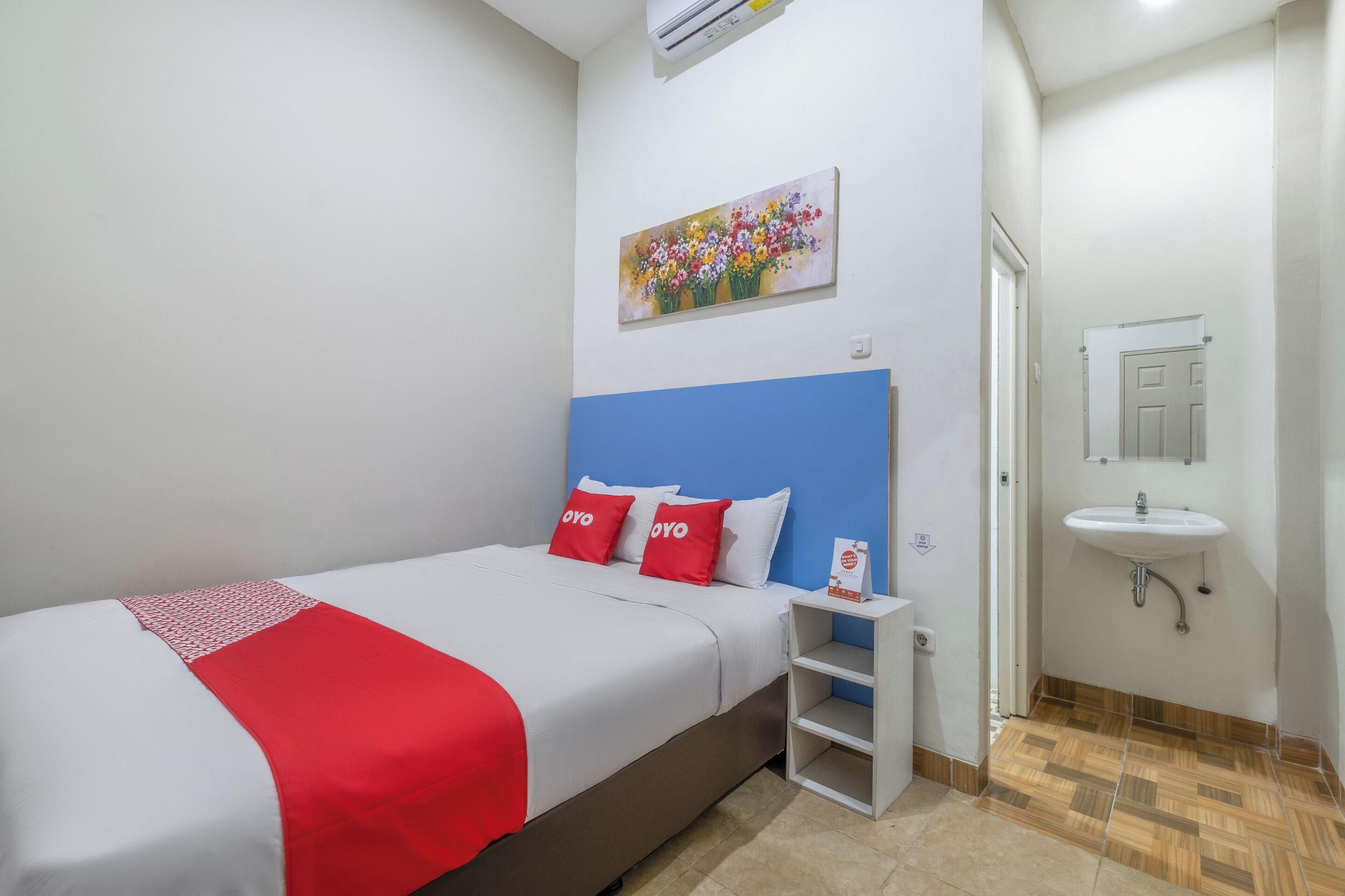 Une chambre d’hôtel avec un lit, une petite table de chevet, un climatiseur fixé au mur, un tableau, une salle de bain avec un lavabo et une porte.