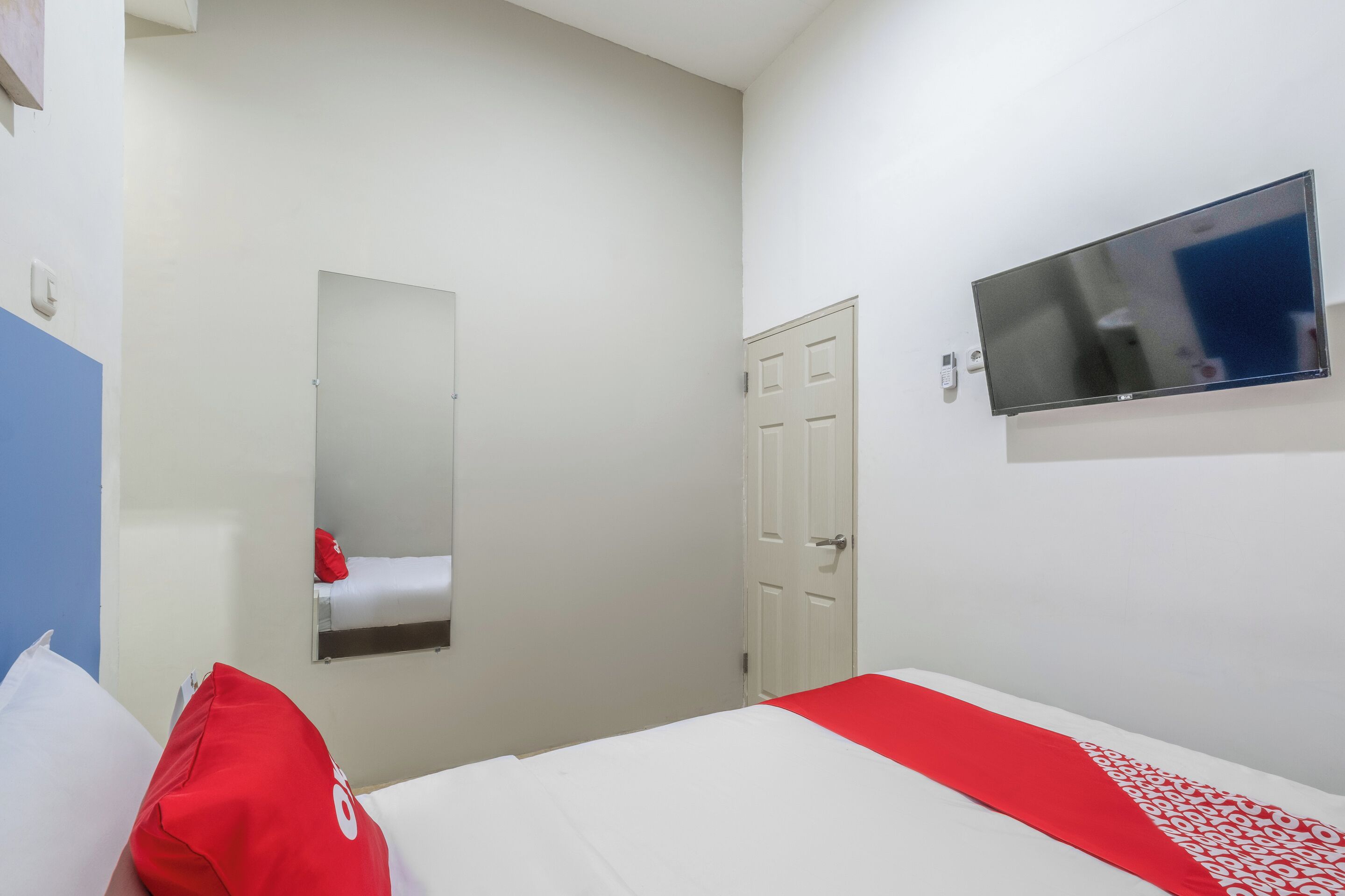 A hotel room with a bed, a red and white bedspread, a flat-screen TV mounted on the wall, a white door, and a mirror on the wall.