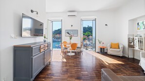 A modern living room with a wooden floor, a grey cabinet, a TV, a dining area with chairs, and a sofa with a yellow pillow. - Sunlight Properties - Capitaine 54 - 1 Bed rental apartment, balcony, port view (Nice)