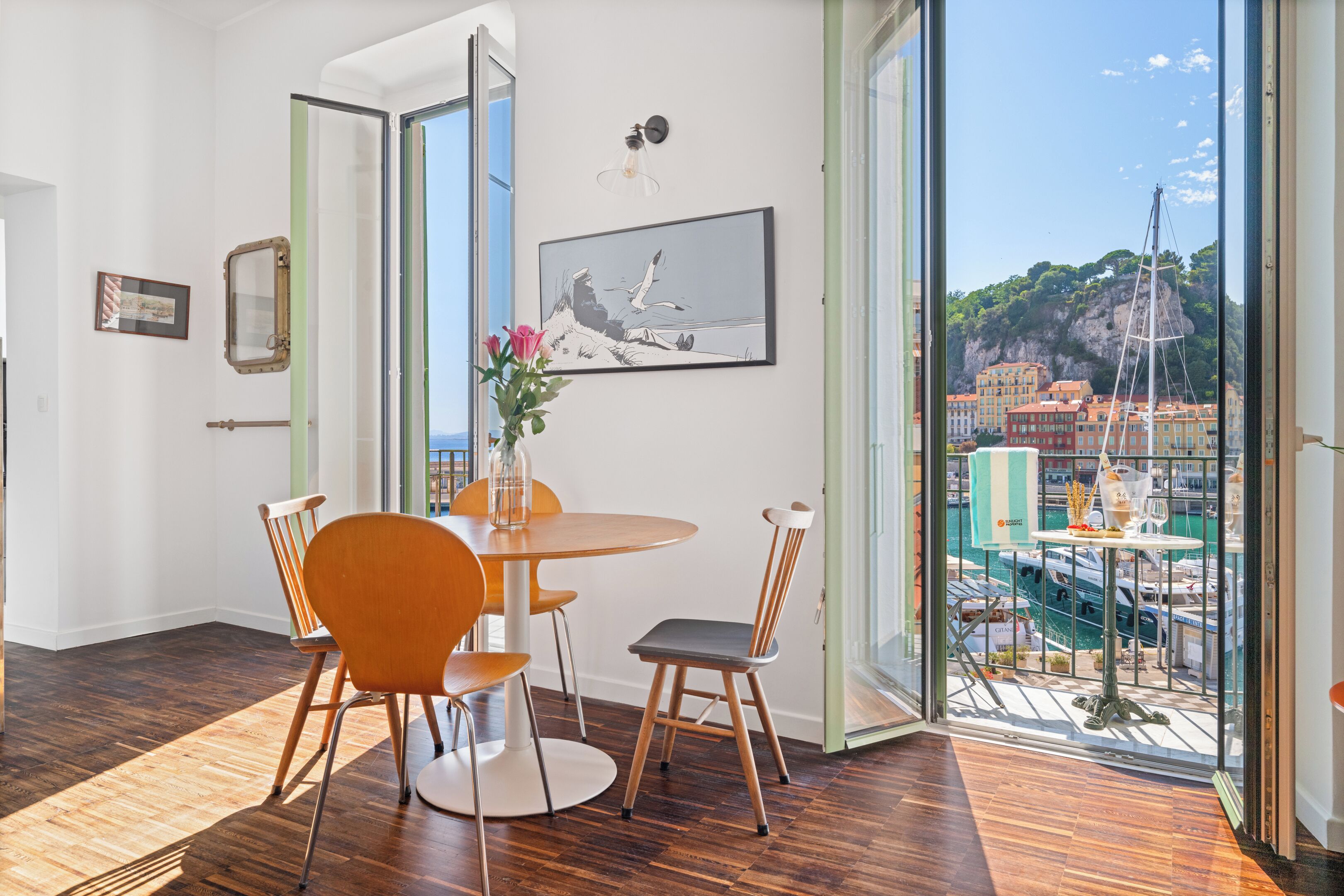 A dining area with a round table and chairs, a vase with flowers, a framed picture, and a view of a coastal town through large windows.