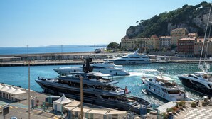 A marina with several yachts docked, a harbor with buildings, and a mountainous backdrop. - Sunlight Properties - Capitaine 54 - 1 Bed rental apartment, balcony, port view (Nice)
